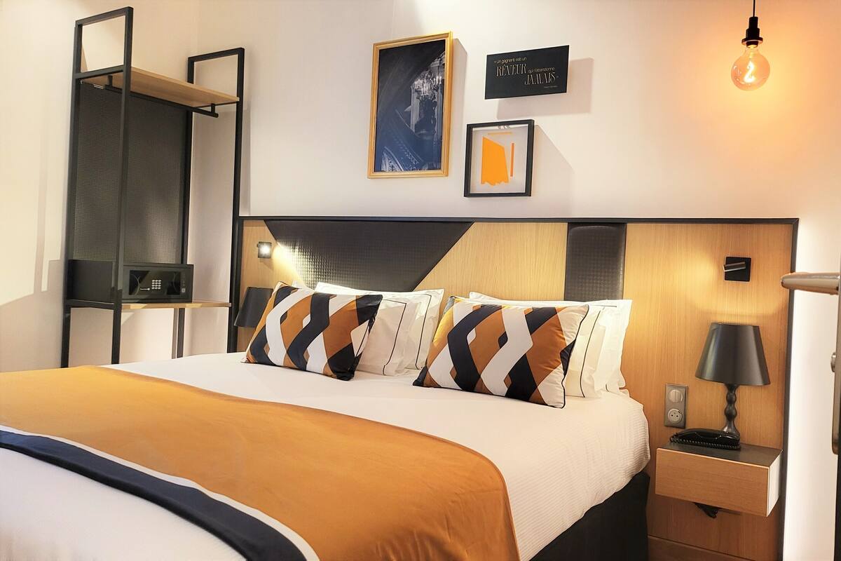 Habitación de hotel con cama, mesita de noche con lámpara, estante en la pared y cuadros enmarcados.