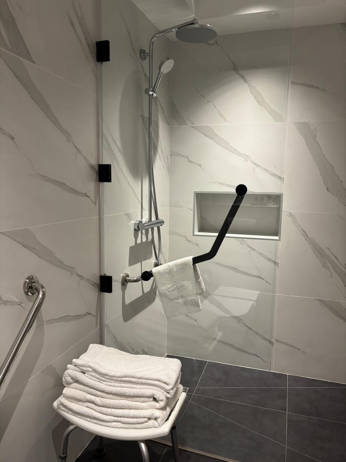 A modern shower with a handheld showerhead, a handheld scrubber, and a towel rack with a folded towel.