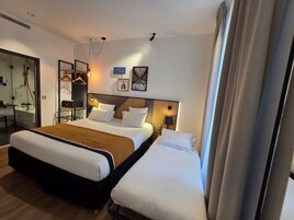 Una camera d'albergo con un letto grande, un letto più piccolo, una scrivania con una sedia, uno specchio e un angolo doccia.