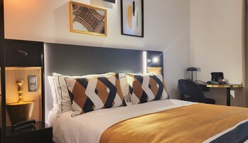 A hotel room with a bed, bedside table, lamp, chair, desk, and artwork on the walls.