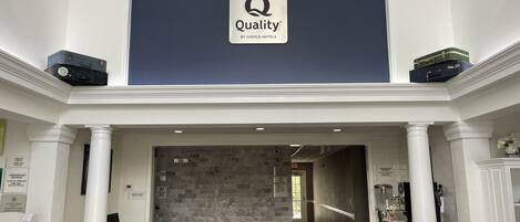 Un vestíbulo de hotel con una pared azul oscuro, un letrero que dice "Quality by Choice Hotels" y un mostrador de recepción con una computadora y un cesto de basura.