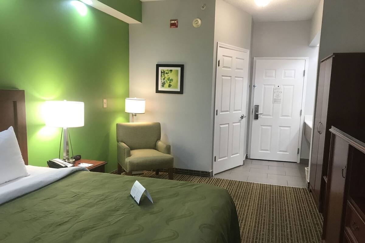 Una habitación de hotel con una cama, una silla, un escritorio con lámpara, un cuadro en la pared y dos puertas.