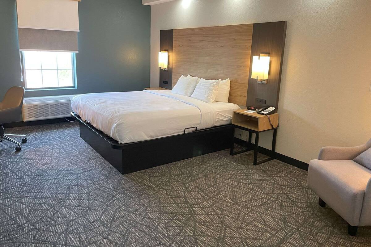 Habitación de hotel con cama, mesita de noche con teléfono, una silla, un escritorio y una ventana con persianas.