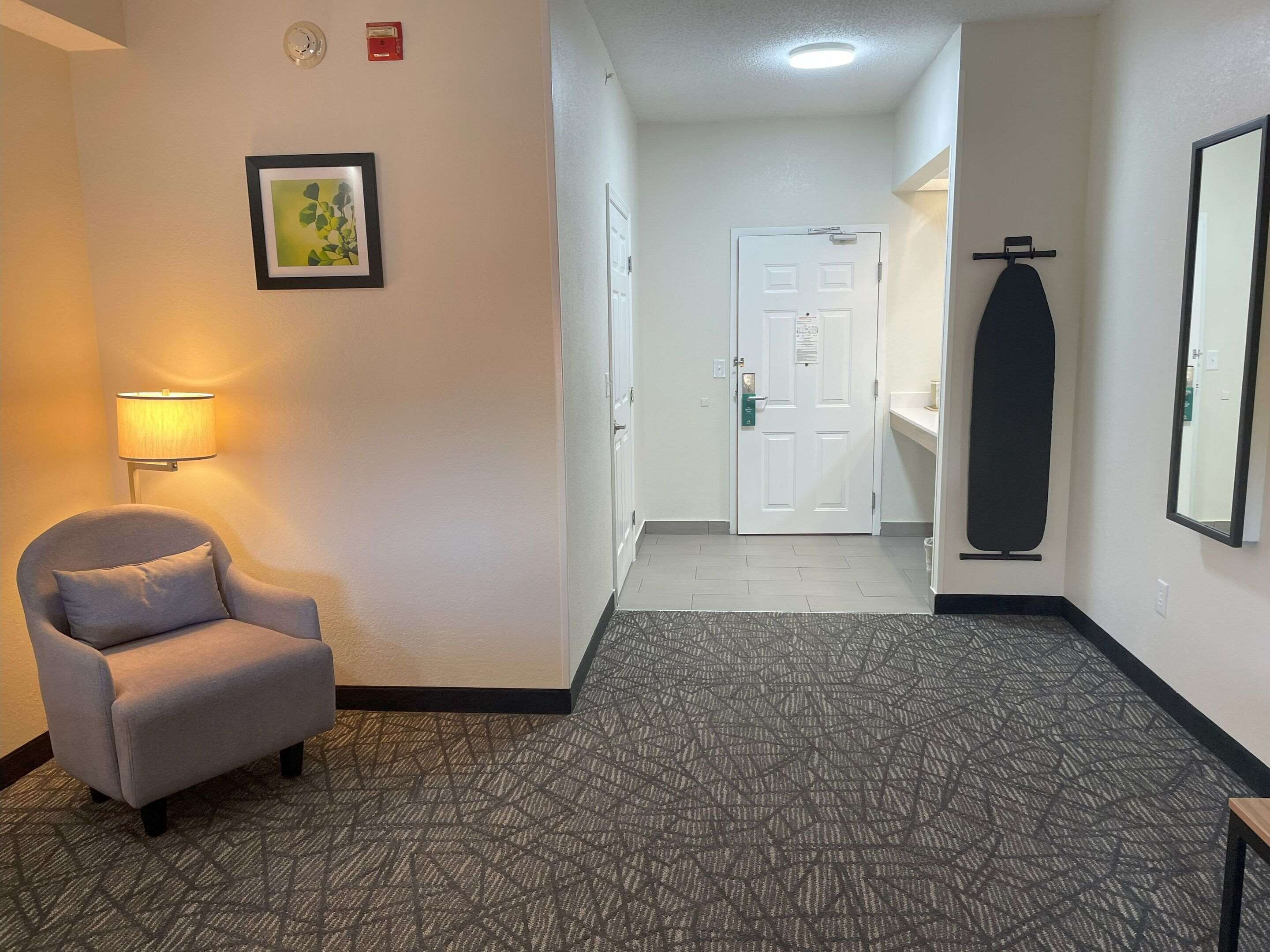 A hotel hallway with a chair, a framed picture, a door, and a mirror.