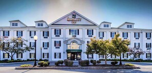 A large, white hotel with a prominent "Quality Inn" sign on the roof, multiple windows, and a well-maintained front garden with trimmed bushes and trees.
