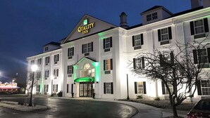 Un grand bâtiment blanc, sur lequel on peut lire l’enseigne « Quality Inn », avec une entrée imposante surmontée d’un auvent éclairé de couleur verte.