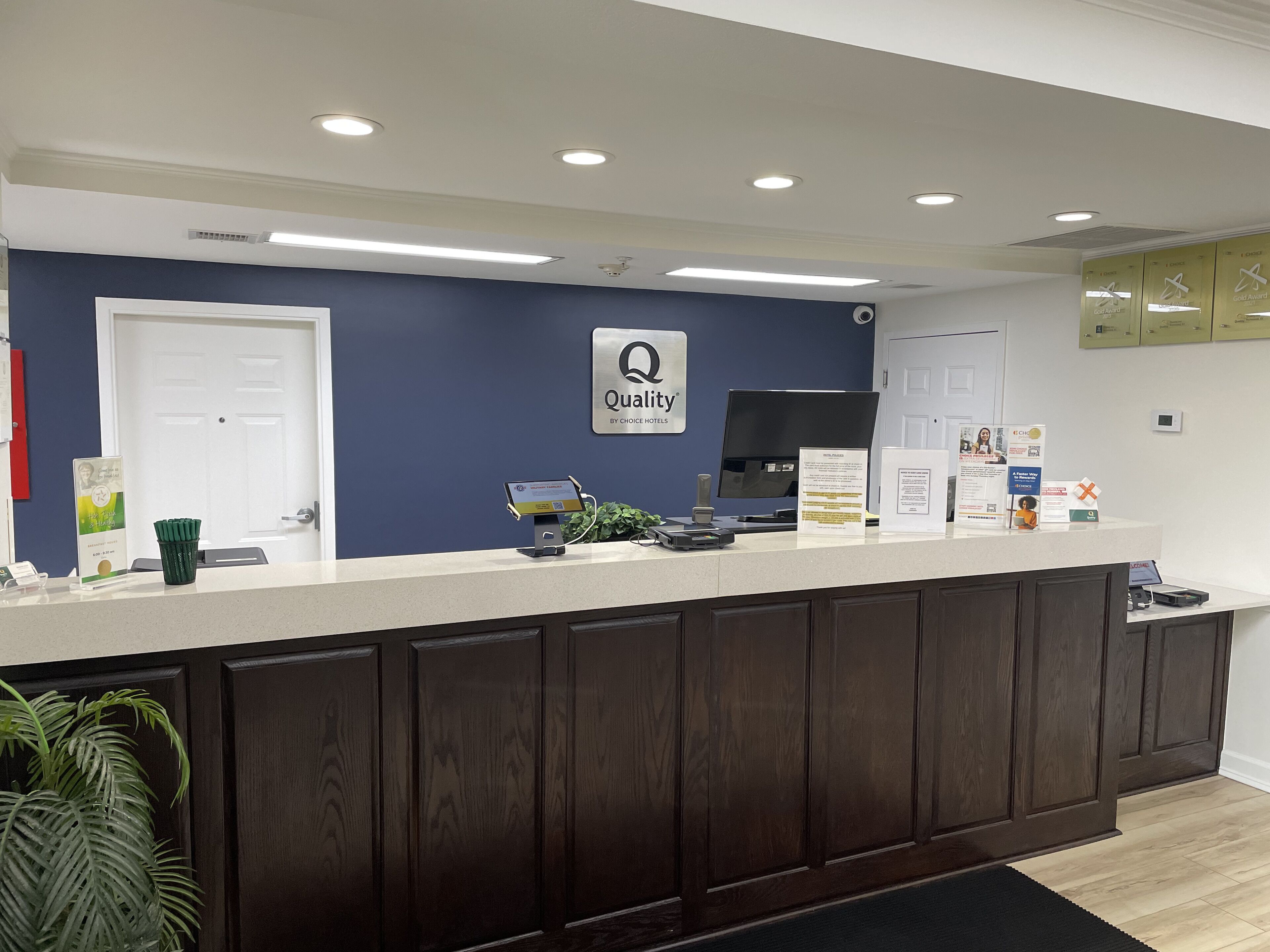 Un mostrador de recepción con un cartel que dice "Quality by Choice Hotels", un monitor de computadora, una planta y folletos informativos.