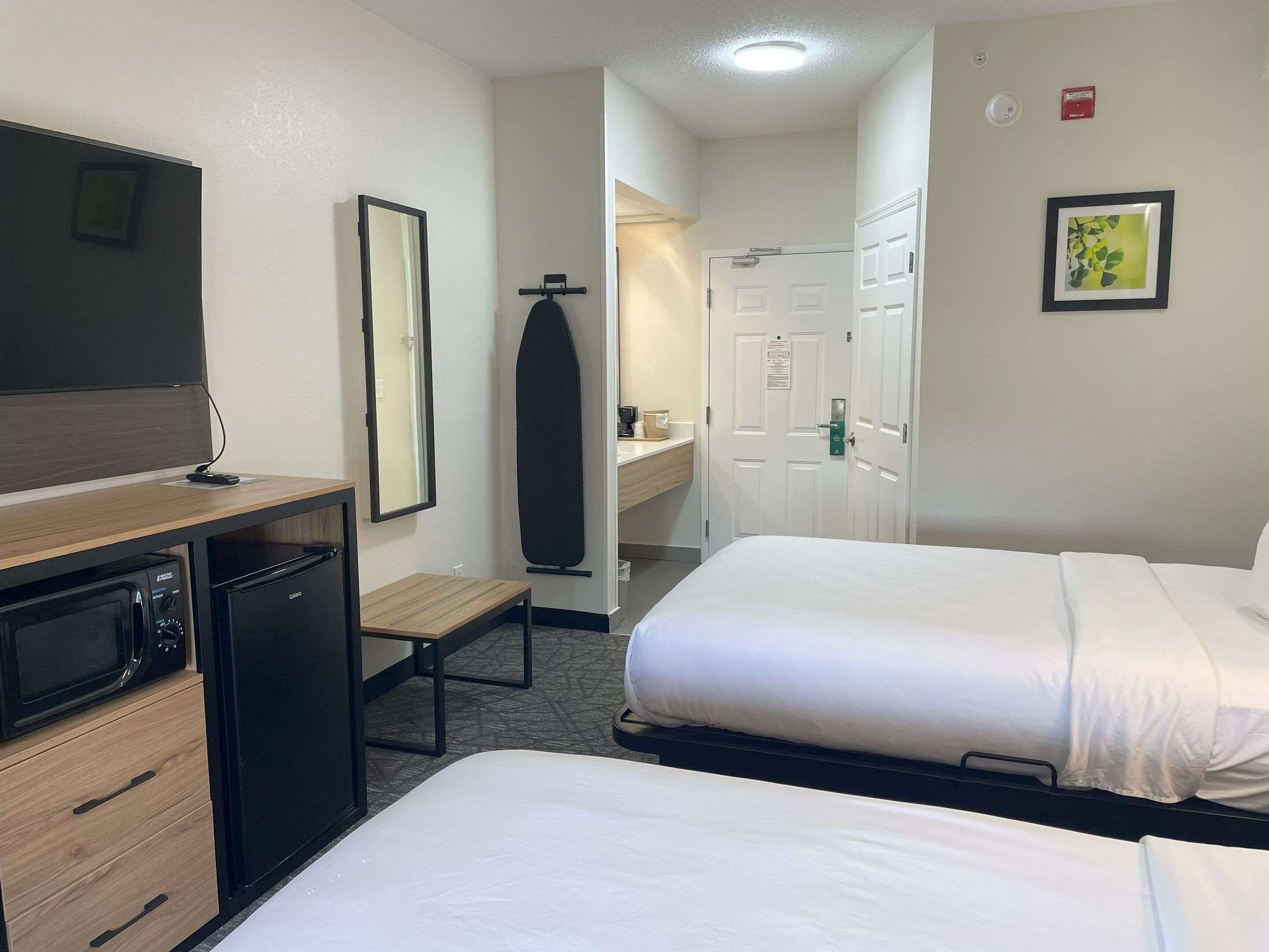Ein Hotelzimmer mit zwei Betten, einem Schreibtisch mit Mikrowelle, einem kleinen Tisch, einem Spiegel und einer Tür zu einem Badezimmer.