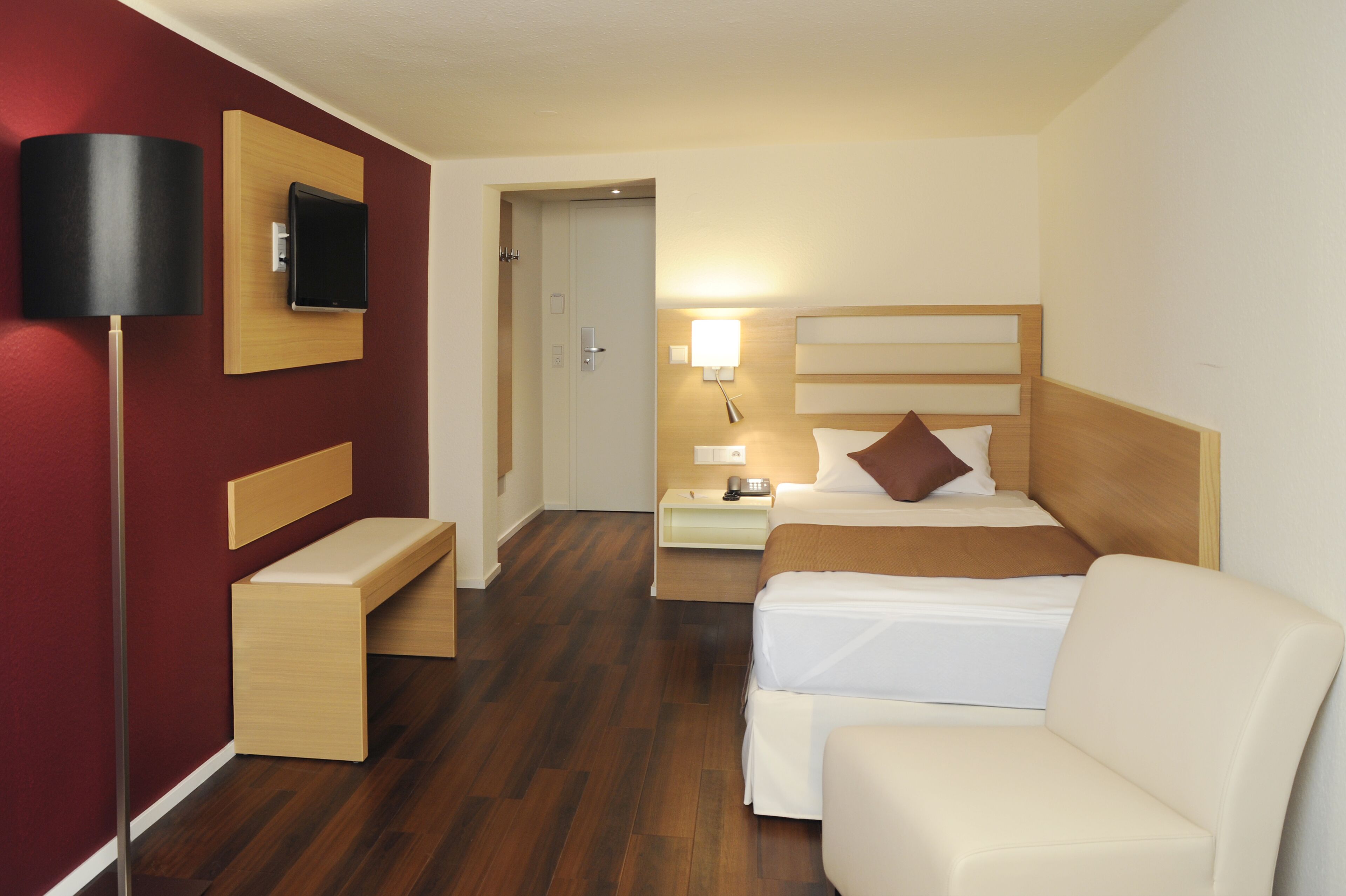 A hotel room with a bed, a desk, a chair, a TV mounted on the wall, and a bedside lamp.