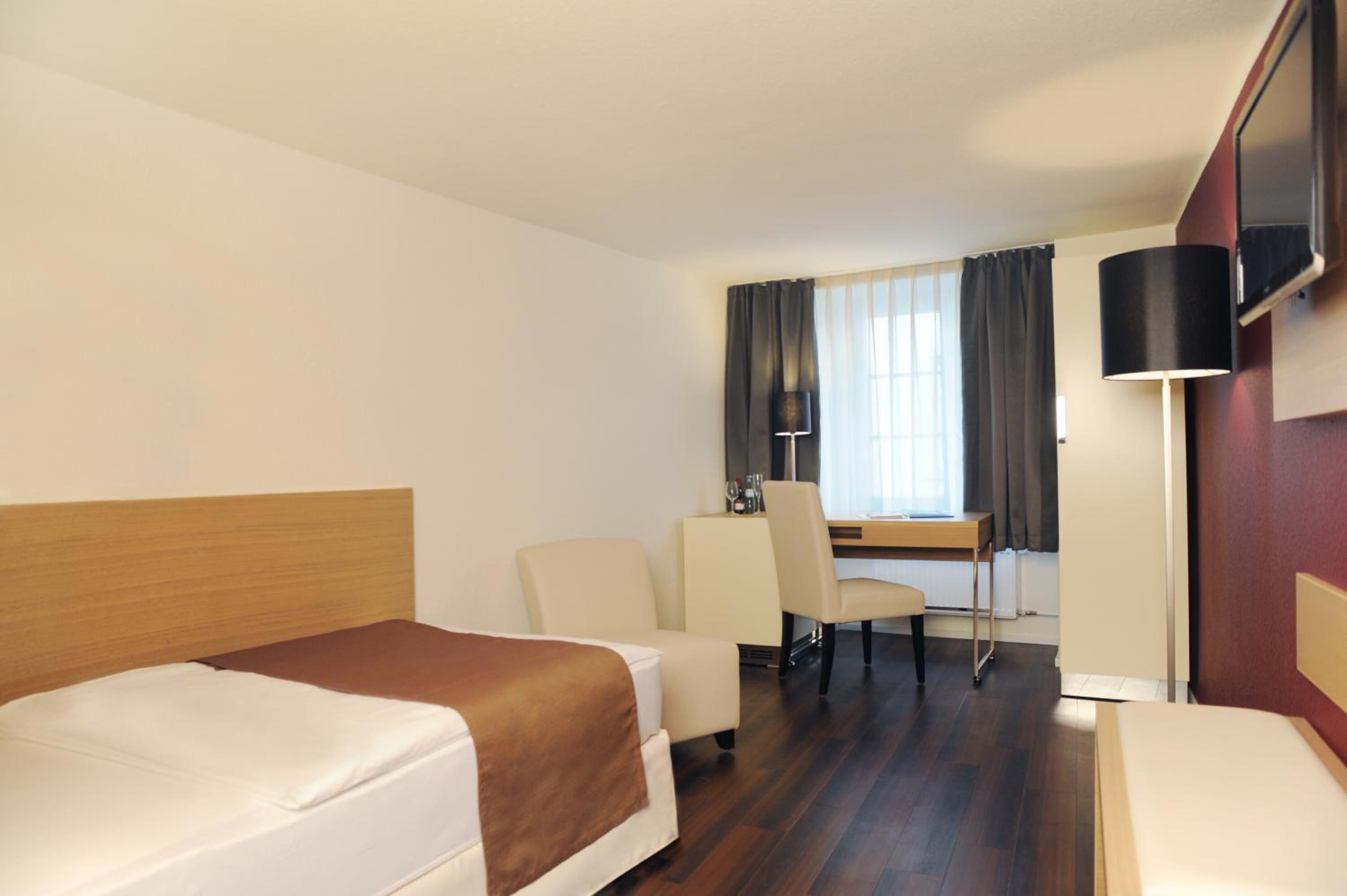 A hotel room with two beds, a desk, a chair, a lamp, and a TV.