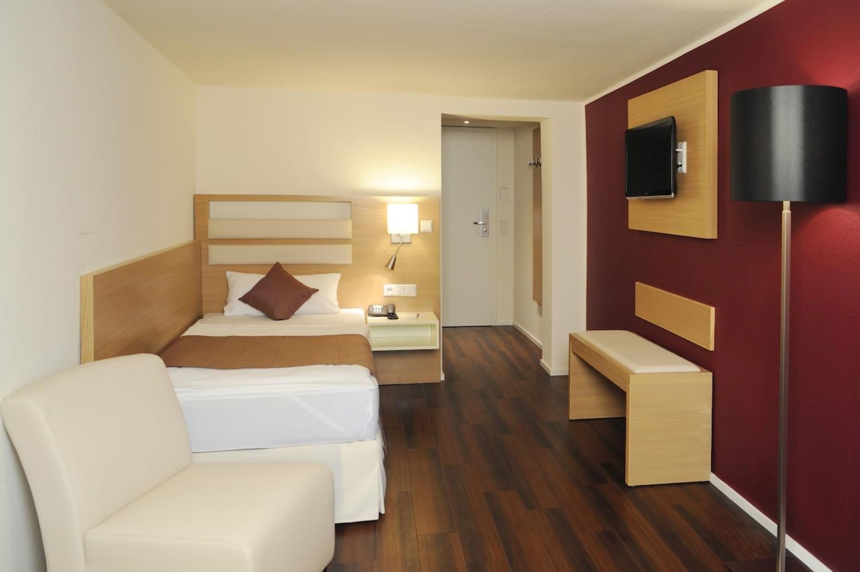 Una habitación de hotel con cama, mesita de noche, lámpara, televisor y un banco.