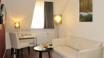 Una habitación de hotel moderna con un sofá blanco, una mesa de comedor con dos sillas, una mesita redonda con una botella y vasos, y un cuadro de un lago.