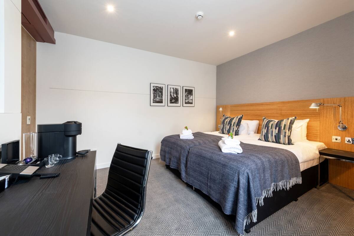 A hotel room with a desk, two beds, a wooden headboard, and a wall-mounted lamp.