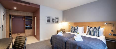 A hotel room with two beds, a desk, a chair, a wardrobe, and a small seating area.