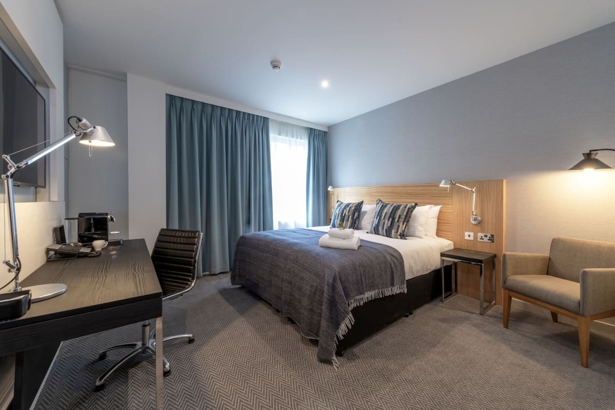 A modern hotel room with a bed, desk, chair, and a small table with a lamp.