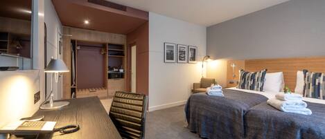 A hotel room with a wooden desk, a bed with a gray bedspread, a headboard, and a nightstand with a lamp.