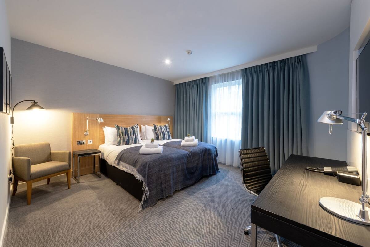 A hotel room with a bed, a desk, a chair, and a lamp.