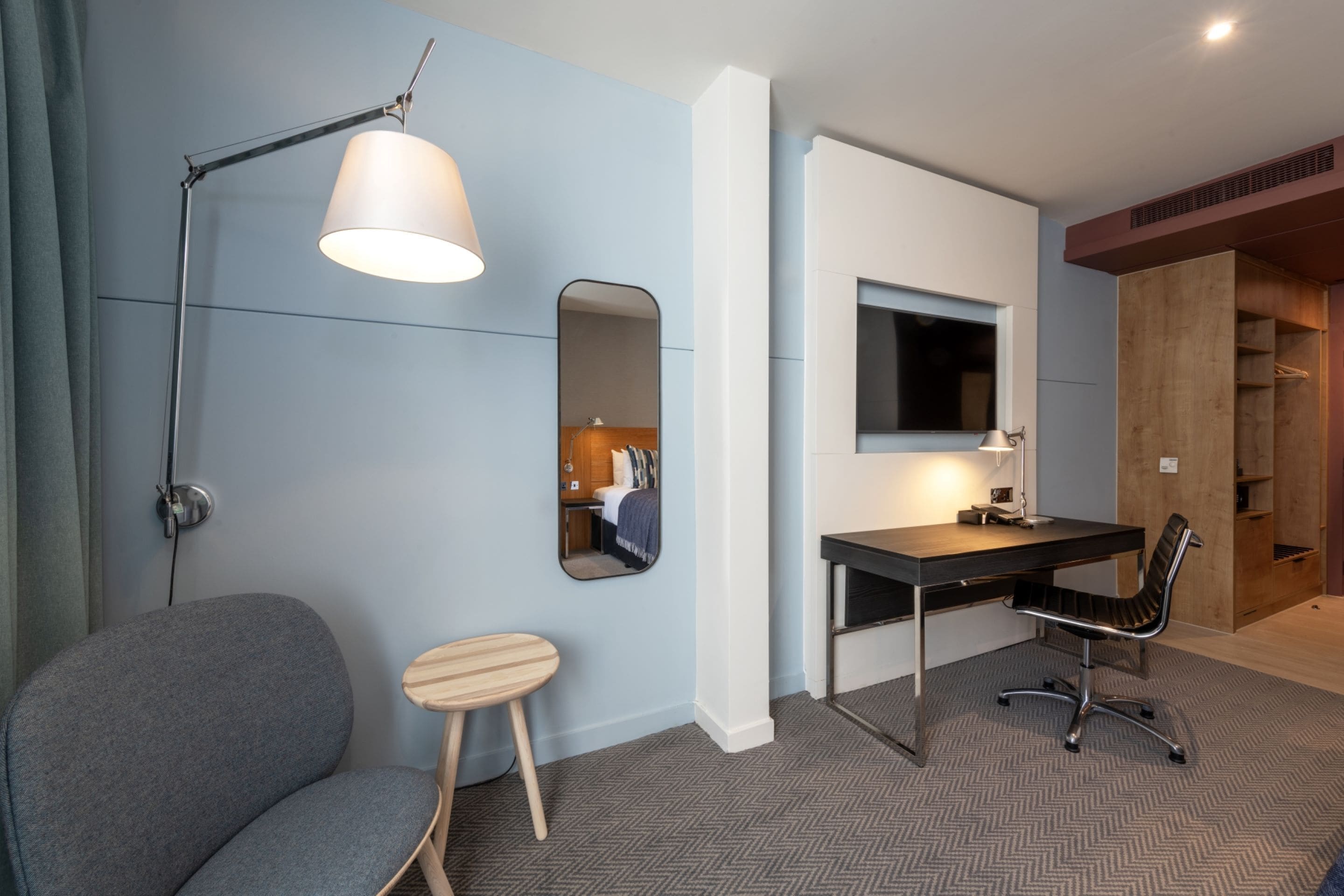 A modern hotel room with a grey armchair, a desk with a chair, a large mirror, and a television mounted on the wall.