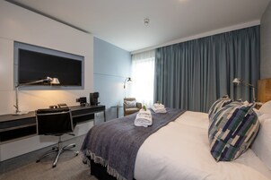 A modern hotel room with a bed, a desk, a chair, a TV, and a window with curtains.