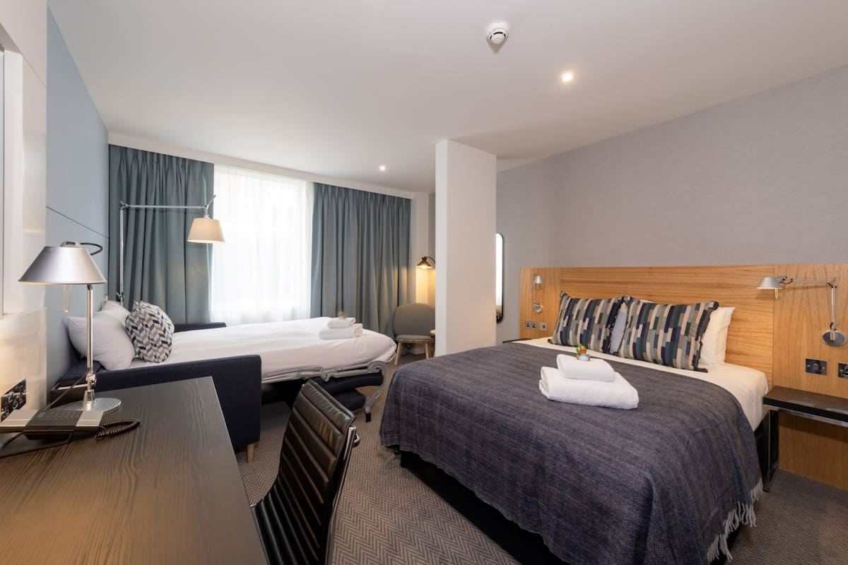 A hotel room with two beds, a desk, a chair, a lamp, and a bedside table.