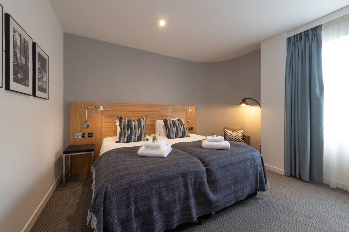A hotel room with two beds, a wooden headboard, a nightstand, a lamp, and a window with curtains.