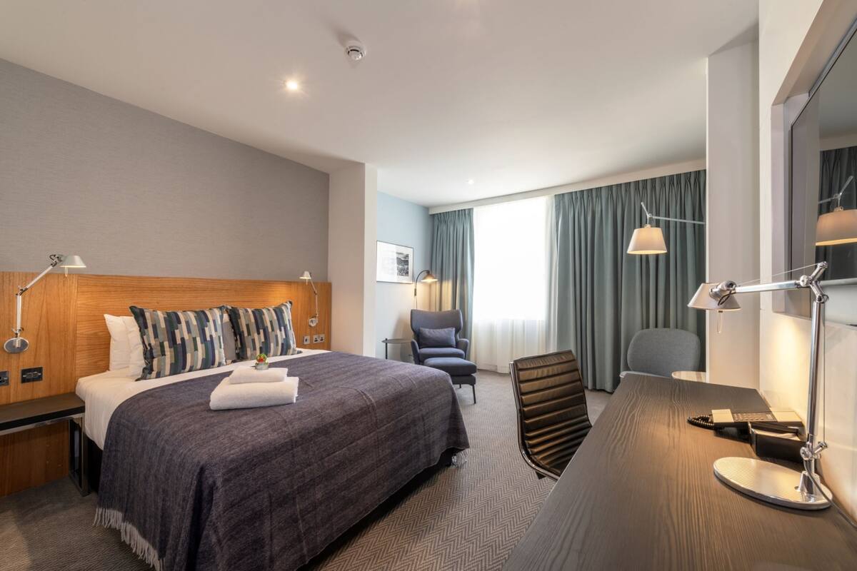 A hotel room with a large bed, a desk, a chair, a lamp, and a window with curtains.