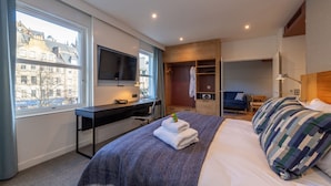 Ett modernt hotellrum med en stor säng, ett skrivbord med en dator, en TV och utsikt över staden genom ett stort fönster.
