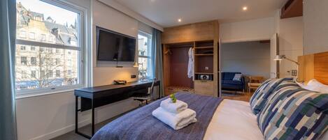 A modern hotel room with a large bed, a desk with a computer, a TV, and a view of the city through a large window.