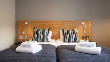 A hotel room with two beds, each with a grey bedspread and white towels. There are two wooden headboards with a camouflage-patterned pillow on each bed.