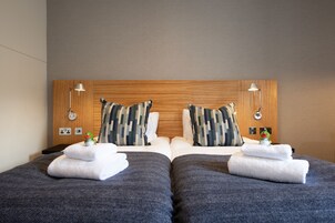 A hotel room with two beds, each with a grey bedspread and white towels. There are two wooden headboards with a camouflage-patterned pillow on each bed.