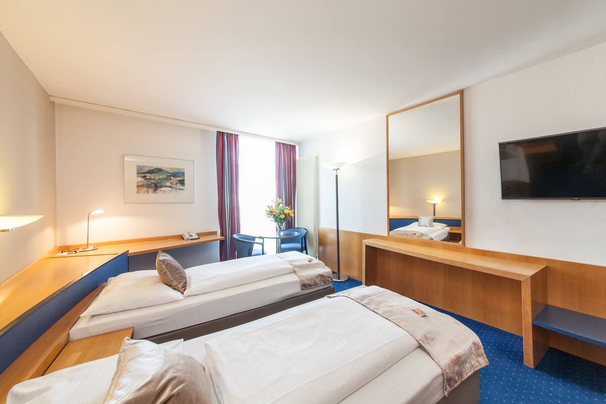 A hotel room with two single beds, a desk, a chair, a TV, and a window with curtains.