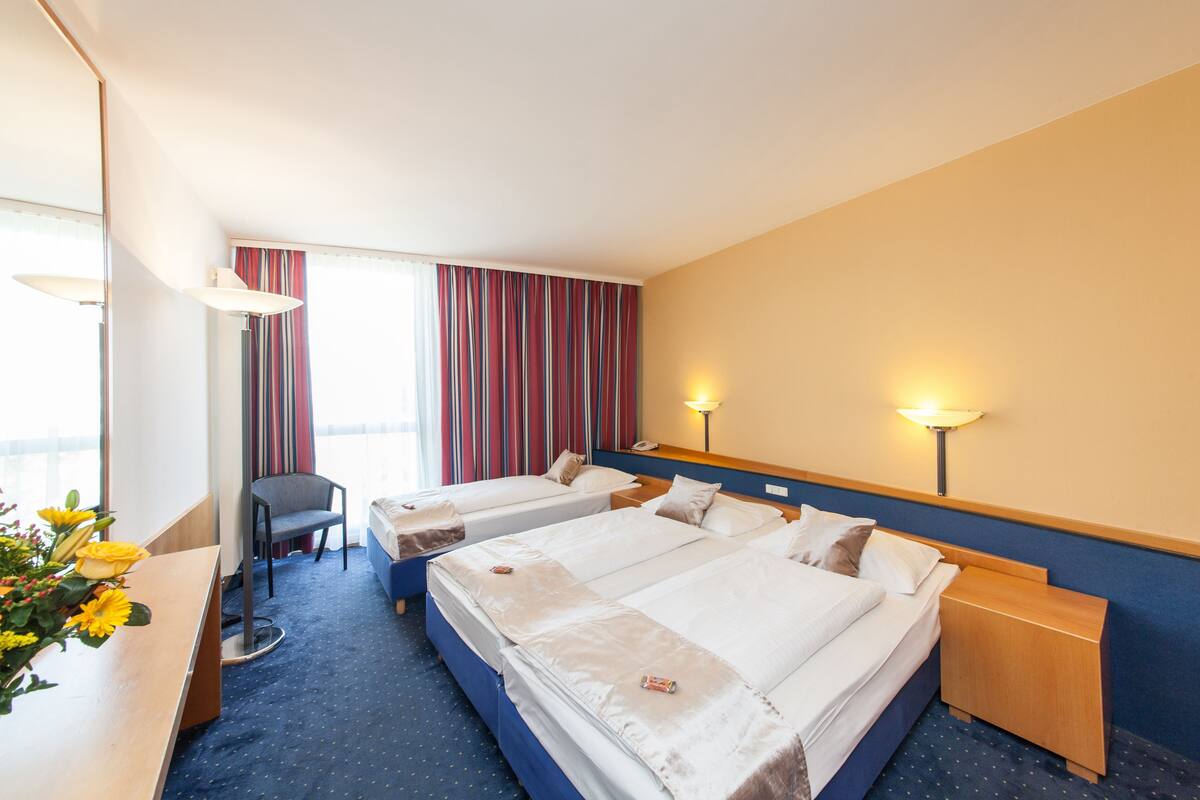 A hotel room with two beds, a desk, a chair, and a window with striped curtains.