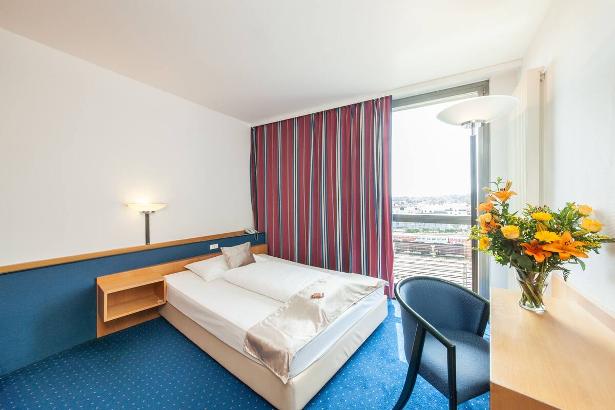 A hotel room with a bed, a desk, a chair, a lamp, and a window with a view.
