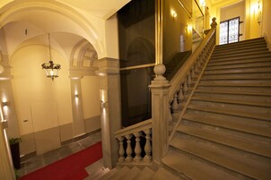 A grand staircase with a red carpet leading to a door, flanked by columns and a hanging light fixture.