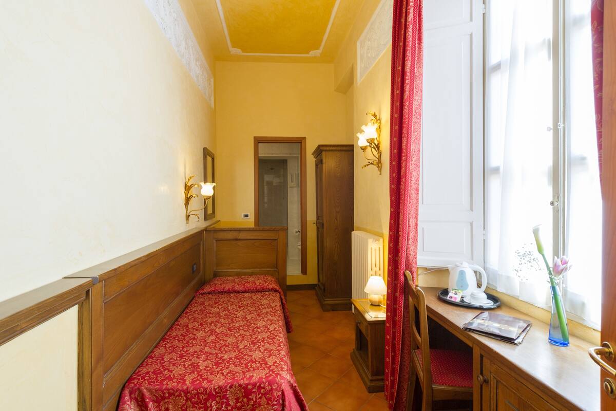 A hotel room with two single beds, a wooden desk with a lamp, a radiator, and a window with curtains.