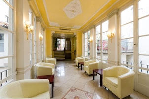 A hotel lobby with a yellow and white color scheme, featuring a central staircase, seating area with chairs, and large windows.