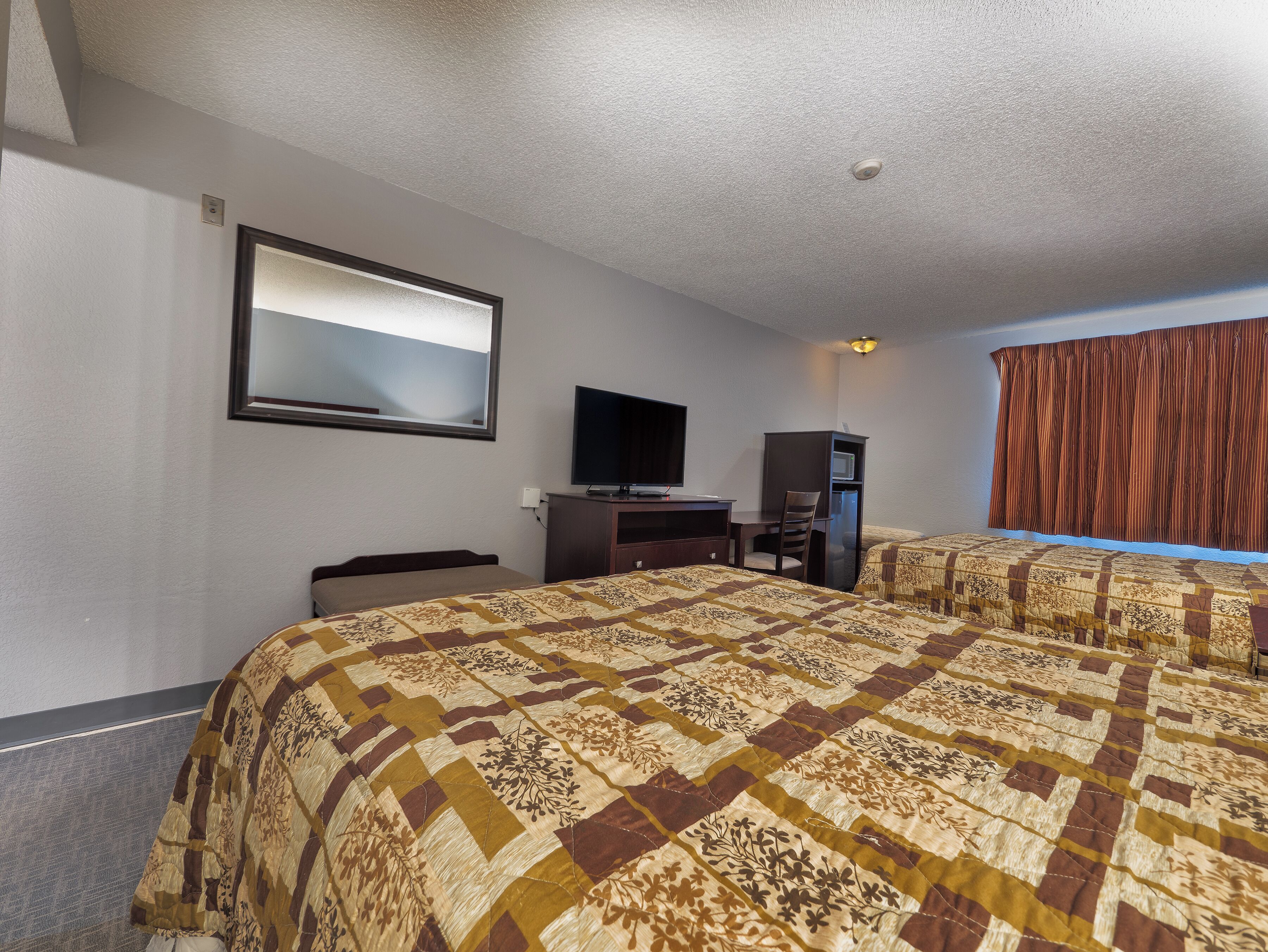 Habitación de hotel con una cama grande, un escritorio con televisor, un mueble pequeño y una ventana con cortinas.