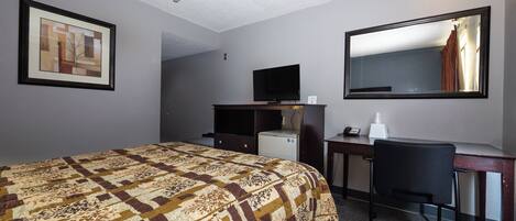 Camera d'albergo con un letto, una scrivania, una sedia, un televisore, un frigorifero e uno specchio.
