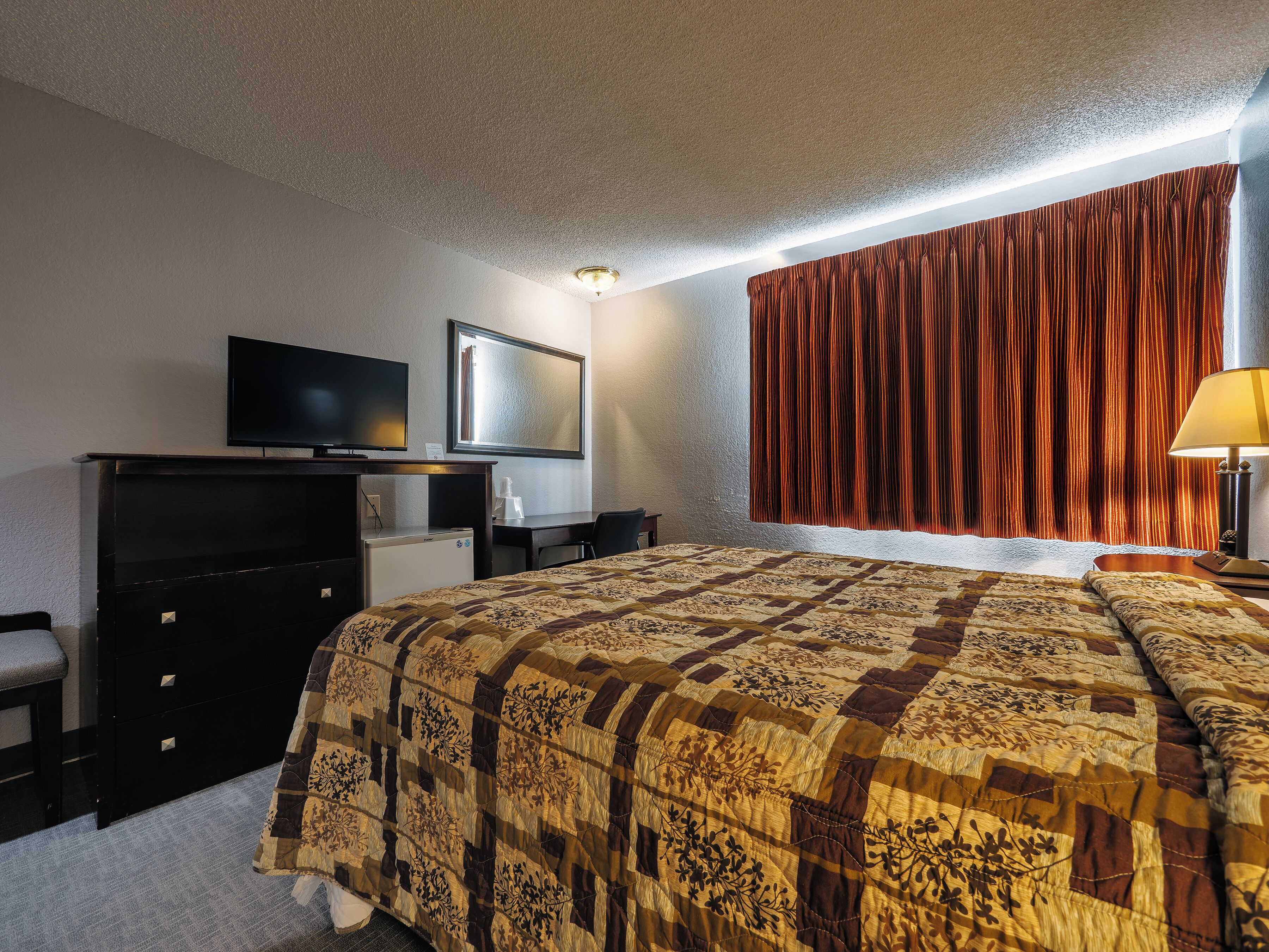 A hotel room with a bed, a desk with a television, a small refrigerator, and a window with curtains.