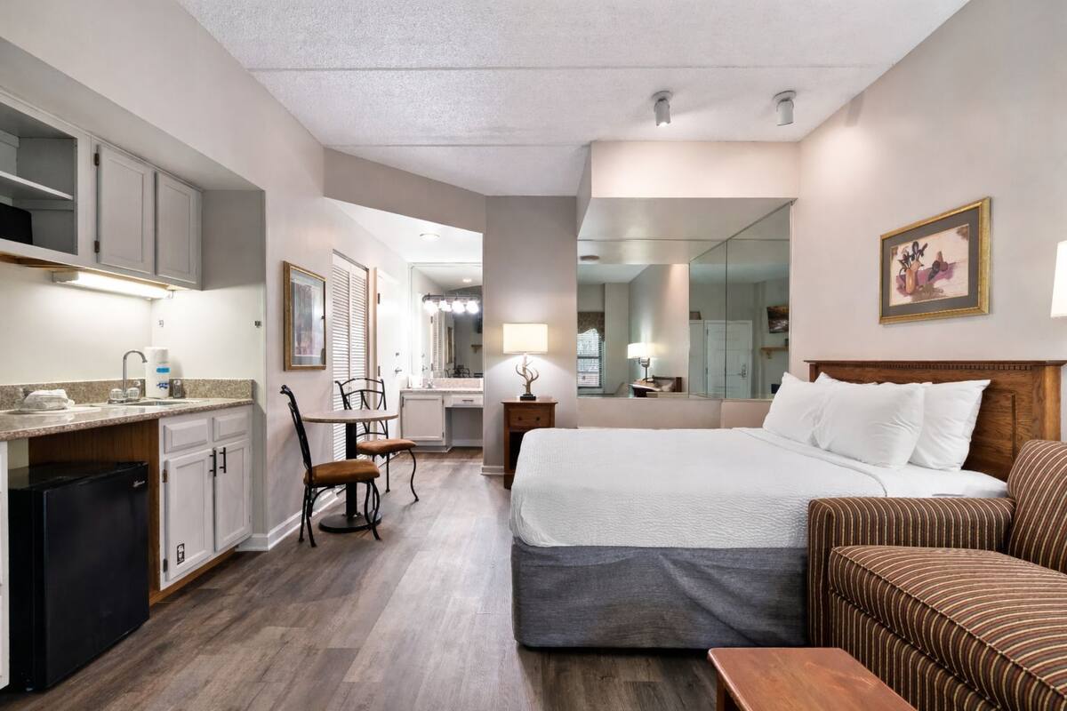 A hotel room with a bed, a sofa, a desk with a chair, a kitchenette, and a bathroom.