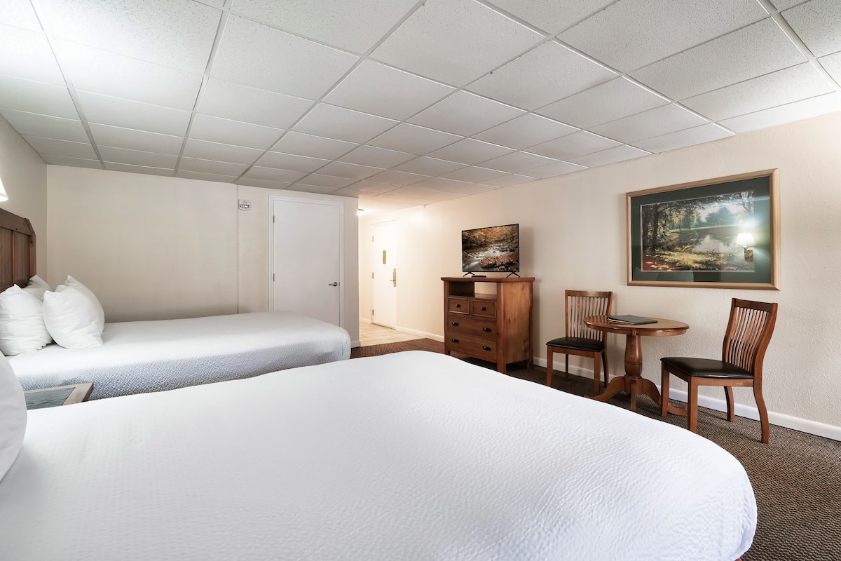 A hotel room with two beds, a wooden dresser, a small table with chairs, and a painting on the wall.
