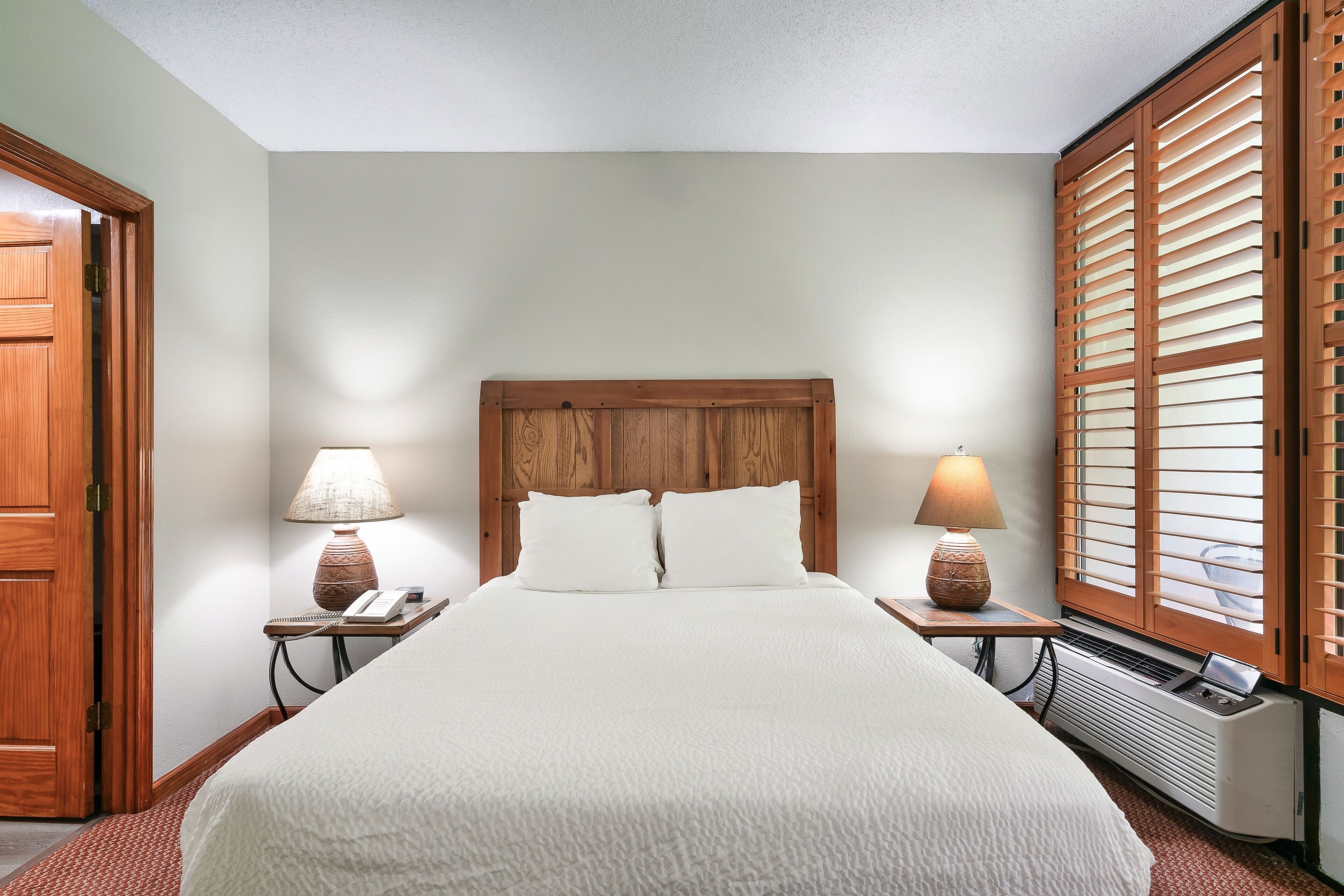 A neatly made bed with a wooden headboard, two bedside lamps, and a window with wooden shutters.