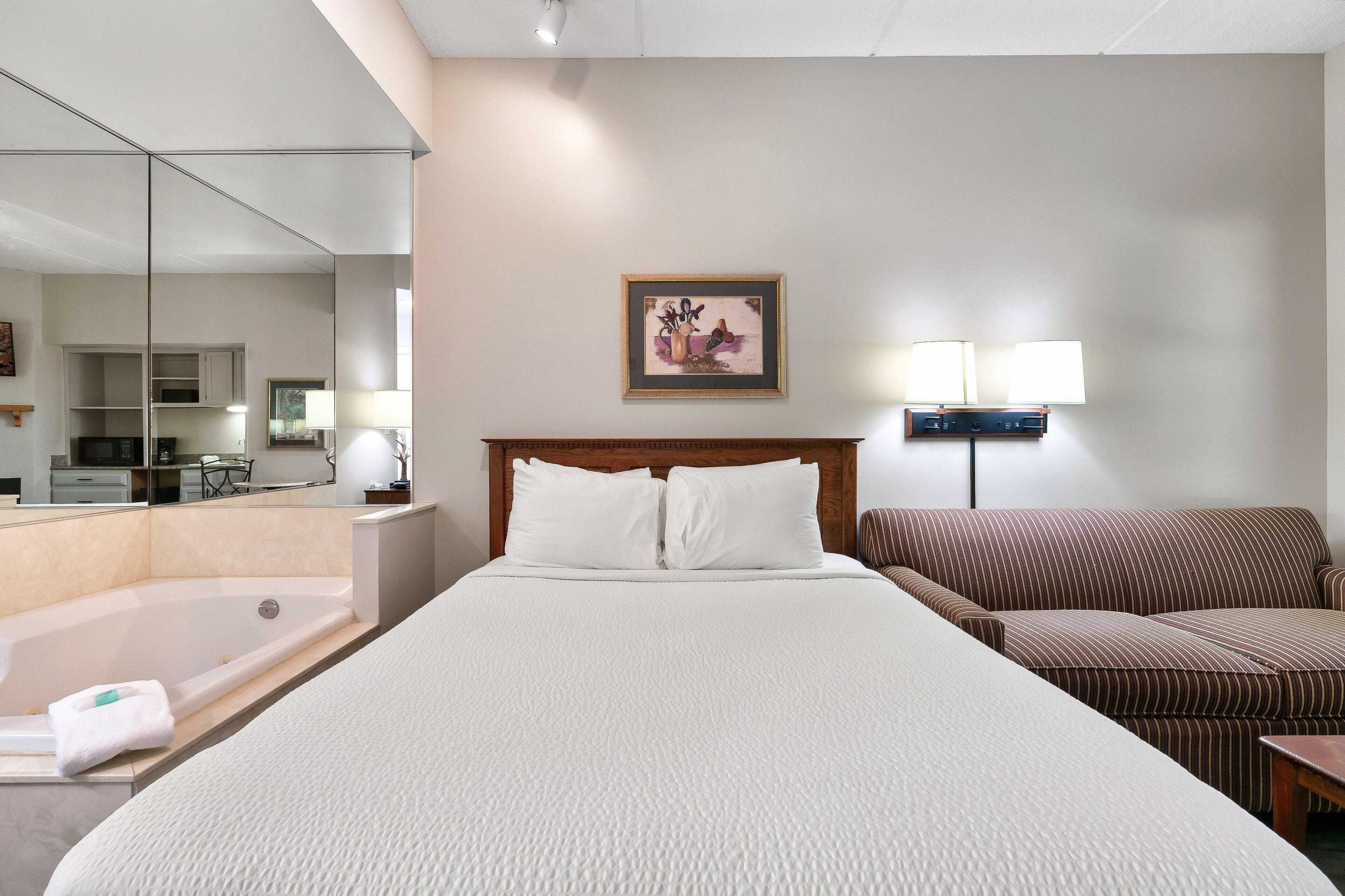 A hotel room with a large bed, a separate sofa, a small table, a lamp, a painting on the wall, and a bathroom with a bathtub and mirror.