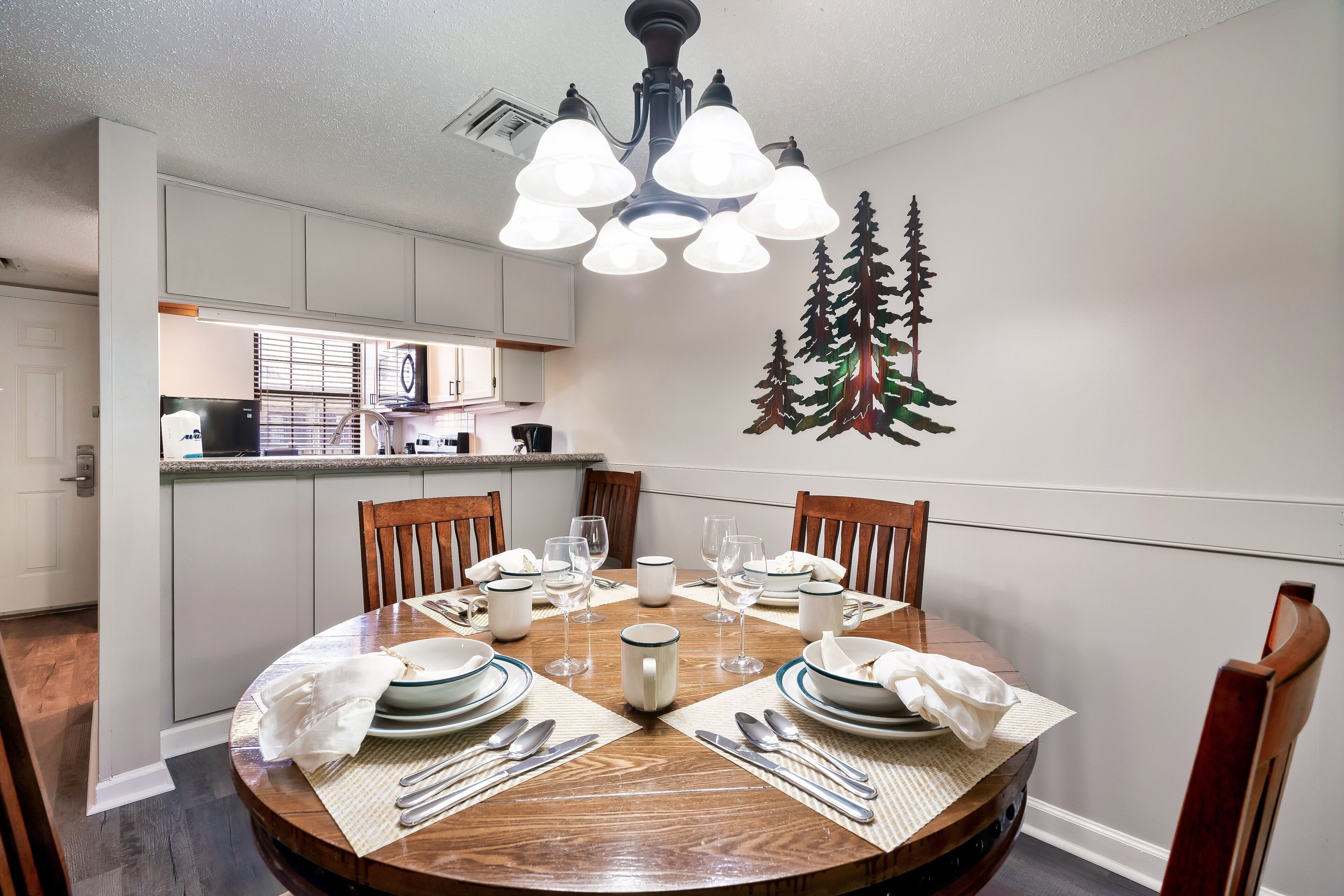 A dining room with a round wooden table set for four, surrounded by chairs. A chandelier hangs above, and a decorative tree is on the wall.