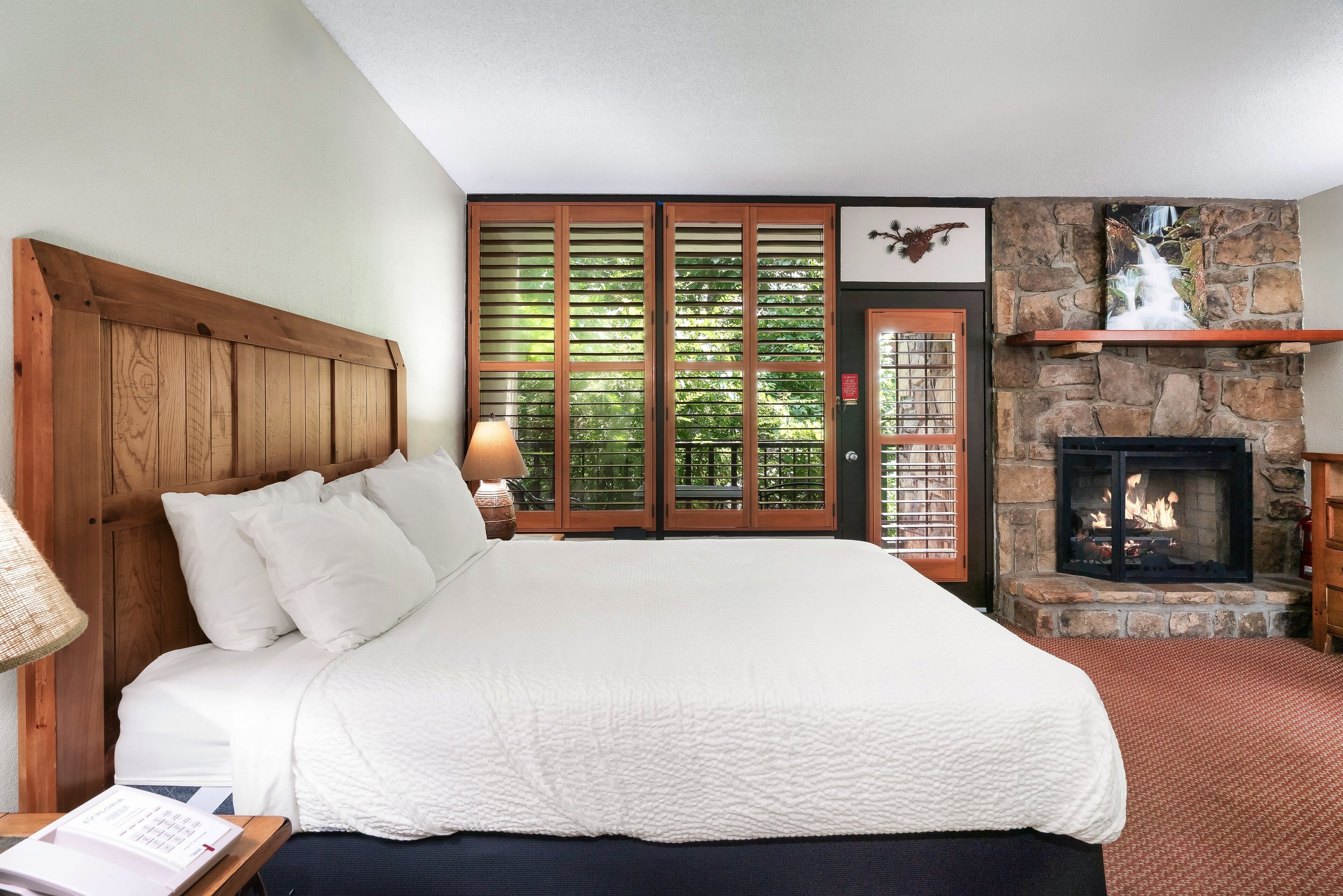 A bedroom with a large bed, a wooden headboard, a stone fireplace, and a view of greenery through a window with wooden shutters.