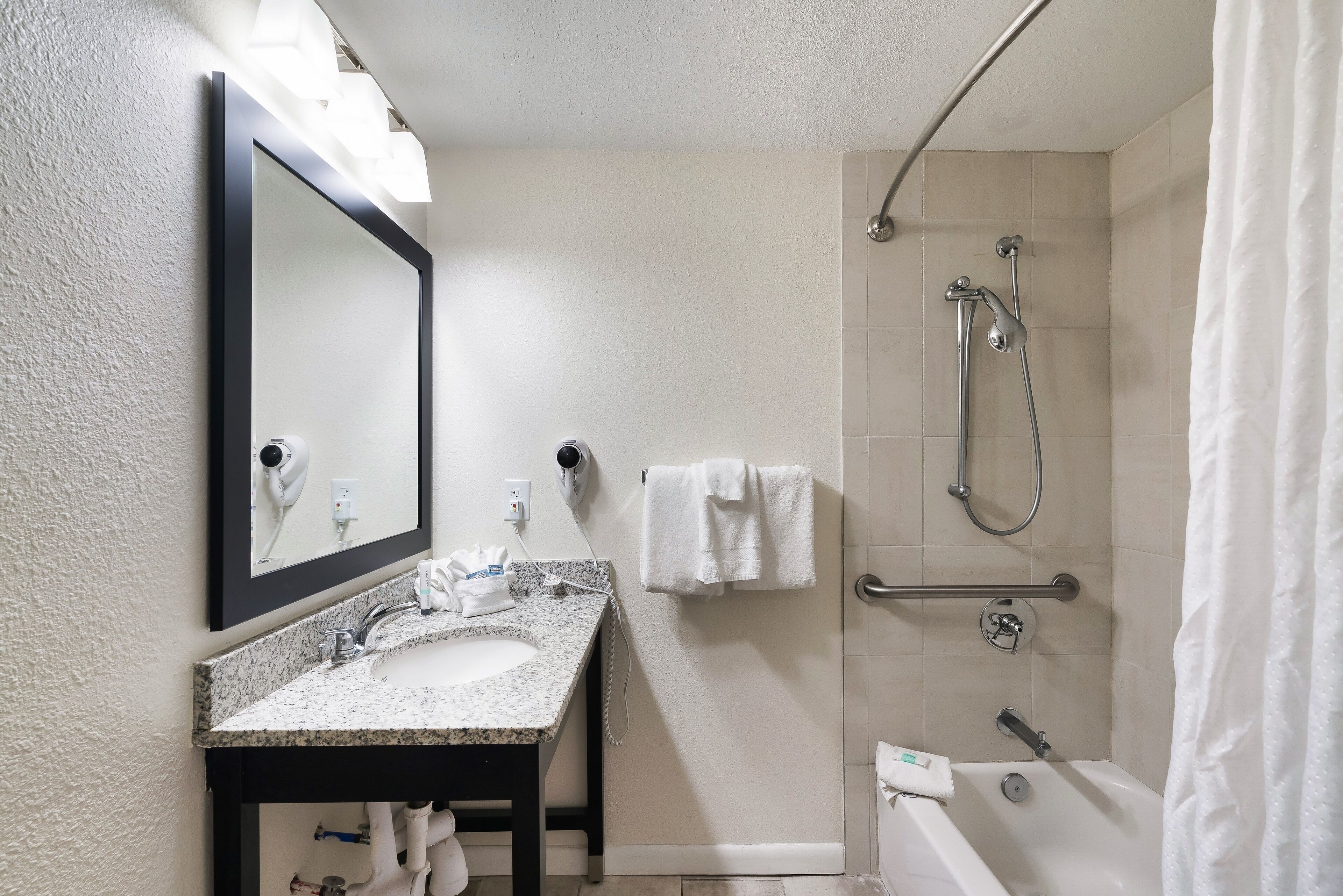 A bathroom with a granite sink, a mirror, a handheld showerhead, a grab bar, and a white bathtub.