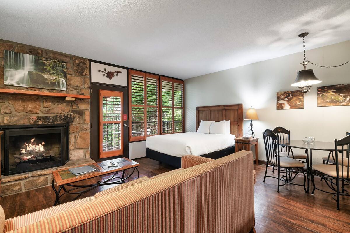 A bedroom with a stone fireplace, a bed, a dining table with chairs, and a sofa.