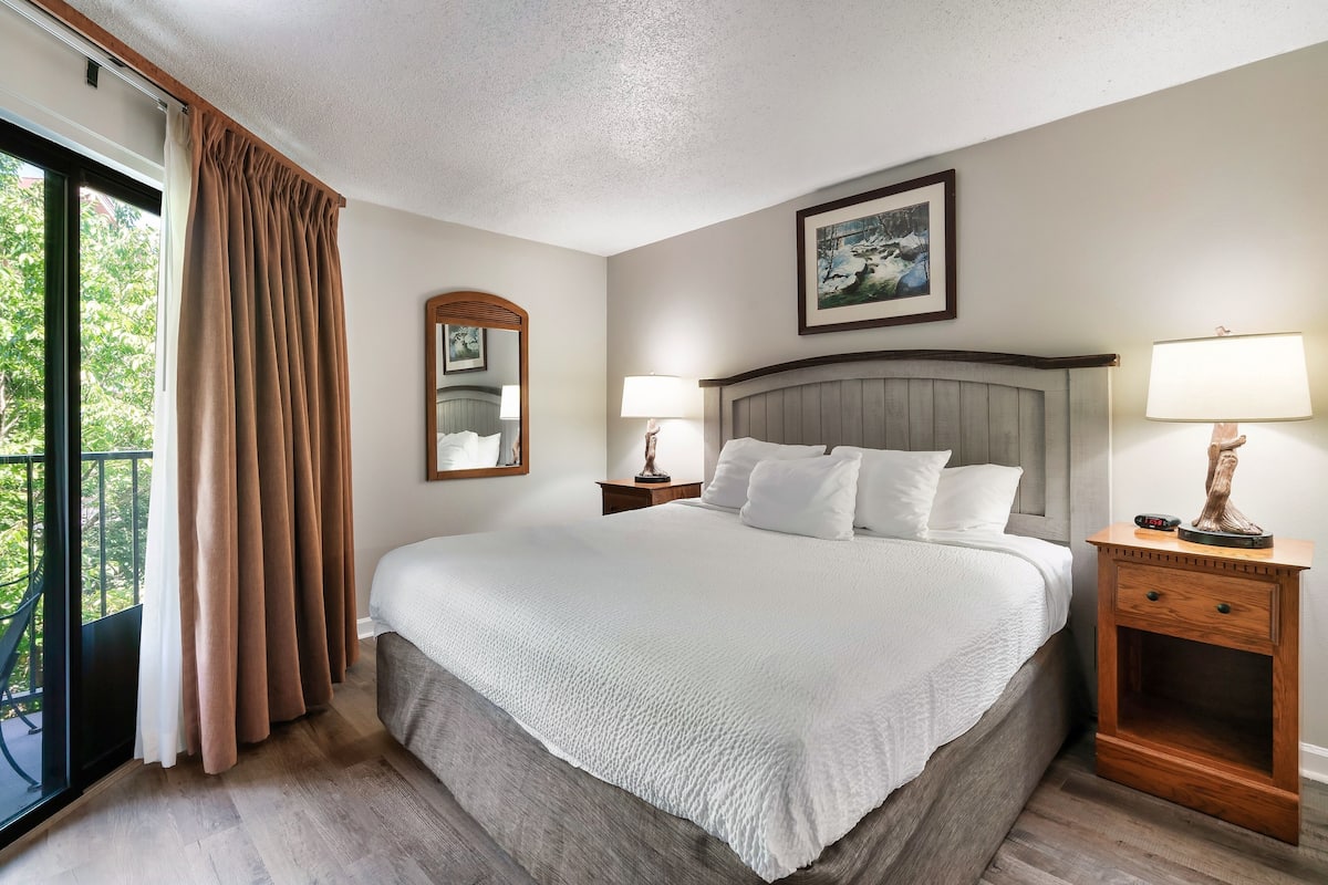A hotel room with a large bed, two bedside tables, a nightstand, a window with curtains, and a framed picture on the wall.