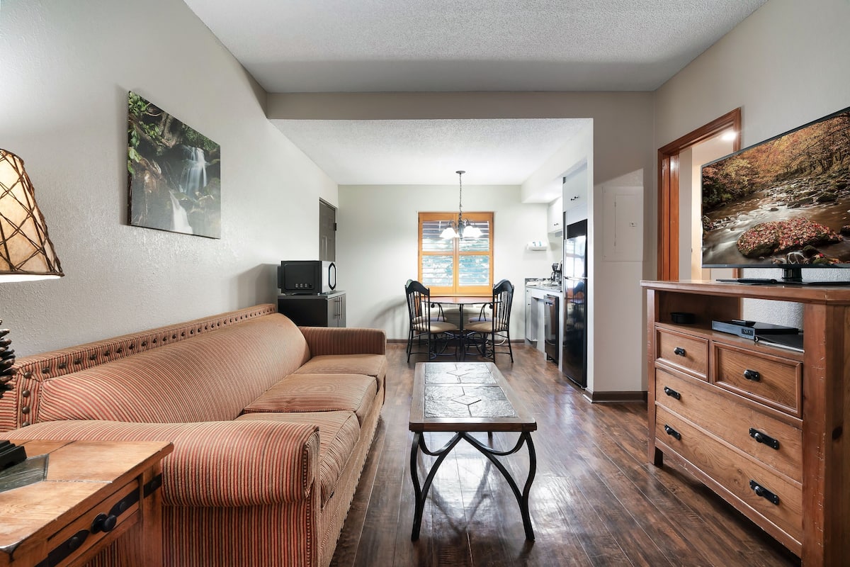 A living room with a sofa, a coffee table, and a TV stand. There is a dining area with a table and chairs.