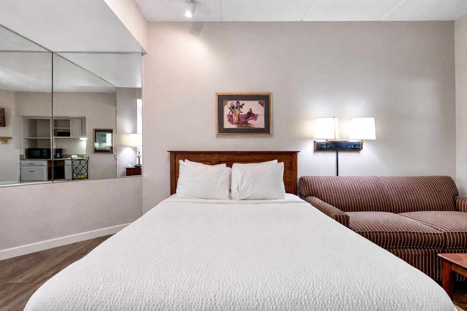 A hotel room with a large bed, a wooden headboard, a bedside table, a lamp, a couch, and a kitchen area visible through a glass partition.